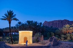 A person in traditional clothing stands before a modern, illuminated structure in an oasis setting, possibly exploring contemporary architecture within a natural landscape.