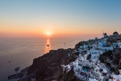 A breathtaking sunset over the Aegean Sea bathes the iconic white-washed buildings of Santorini in a warm, golden light, as sailboats glide across the horizon, creating a picturesque scene of tranquility and beauty.