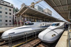 Two Shinkansen bullet trains are parked at a station, with a conductor standing at the door of one of the trains, likely preparing for departure or arrival.