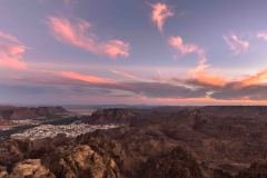 A scenic view of Al-'Ula, Saudi Arabia, at sunset, showcasing the town nestled among the rocky desert landscape under a sky painted with pink and purple hues.