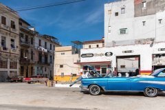A man is working on the engine of a classic blue convertible car in front of a building with a sign that says "Servicentros Oro Negro Zanja Cimex" in Cuba.