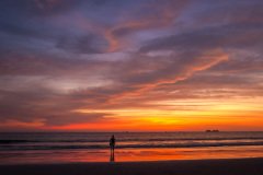 A lone figure stands on the beach, silhouetted against a vibrant sunset, possibly enjoying the tranquility and beauty of the moment.
