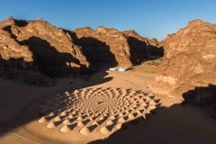 An aerial view shows the desert art installation "The Desert X AlUla 2022" in Saudi Arabia, featuring a series of sand mounds arranged in a spiral pattern, with a building in the background, set against a backdrop of rocky mountains.