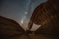 A stunning nightscape captures the Milky Way galaxy arching over a natural rock bridge, showcasing the beauty of the cosmos and the grandeur of geological formations.