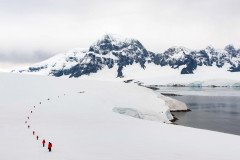 A group of people in red jackets are walking across a snowy landscape in Antarctica, with mountains and water in the background.