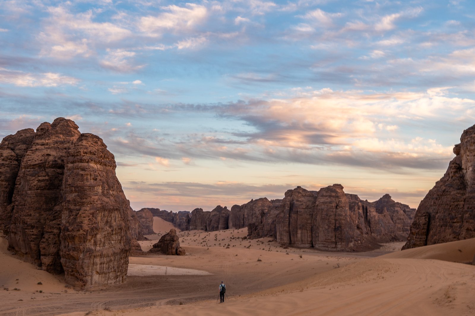 A lone photographer sets up their tripod in the vast desert landscape of AlUla, Saudi Arabia, aiming to capture the beauty of the sandstone formations under the soft hues of the sunset sky.
