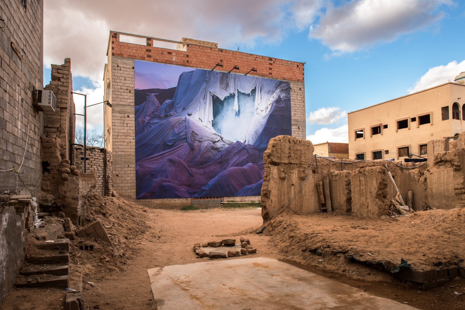 A large mural of draped fabric hangs on the side of a building in a state of disrepair, with rubble and dirt in the foreground.