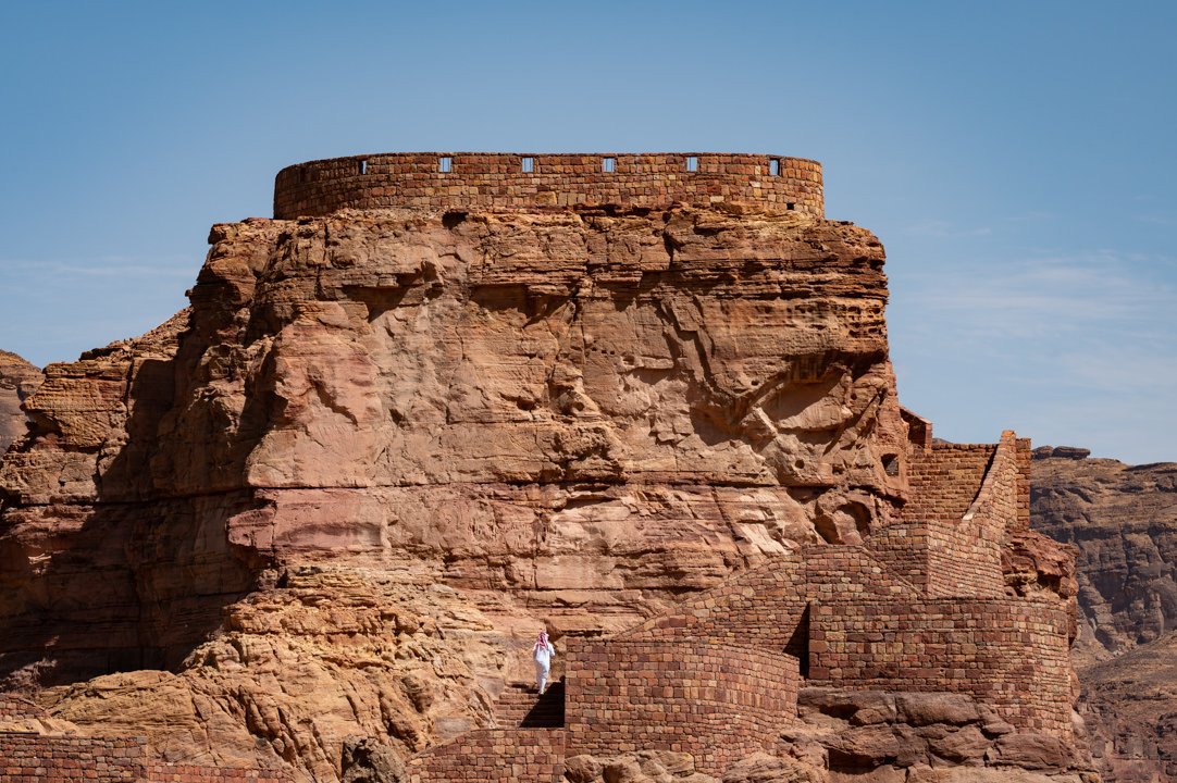 A person in traditional Arab clothing descends the stone steps of a large, ancient, red rock structure, while other people are seen climbing the steps on the other side of the structure, likely exploring the historical site.