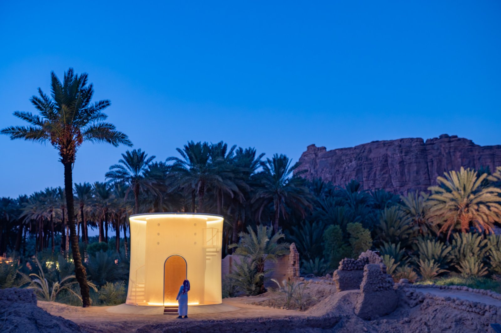 A person in traditional clothing stands before a modern, illuminated structure in an oasis setting, possibly exploring contemporary architecture within a natural landscape.