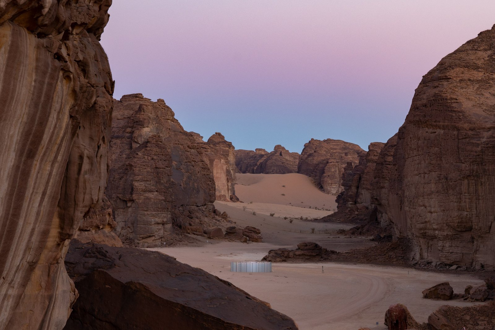A landscape view of a desert valley surrounded by tall rock formations, with a small, modern structure in the foreground, under a pastel-colored sky.