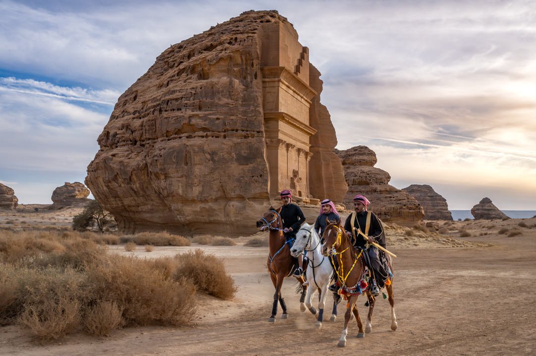 Three men in traditional Saudi Arabian clothing ride horses in front of the Qasr al-Farid tomb in the ancient city of Hegra, also known as Mada'in Salih, a UNESCO World Heritage Site, showcasing the region's rich cultural heritage and history.