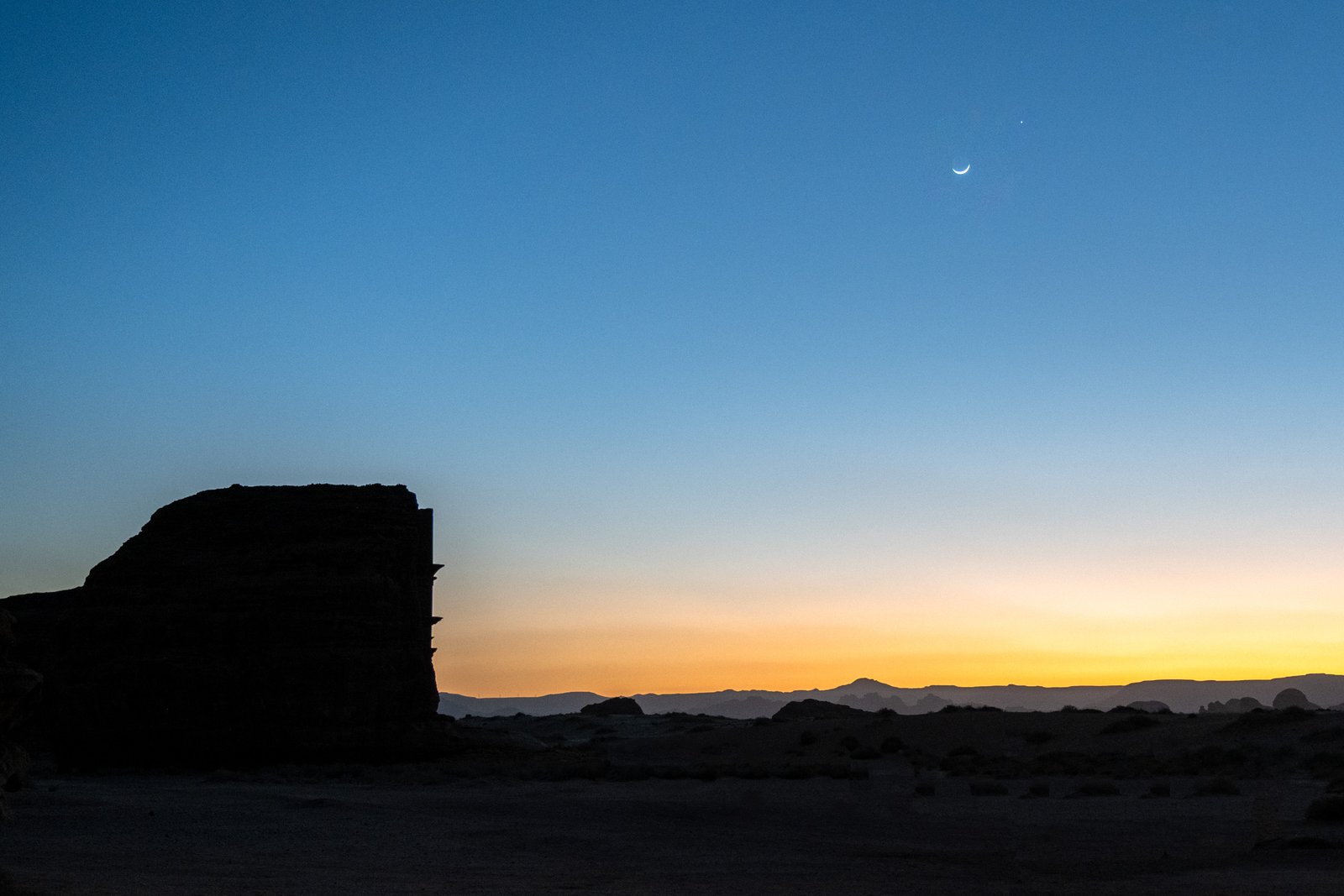 A crescent moon and a star shine in the sky above a desert landscape at sunset, silhouetting rock formations and mountains.